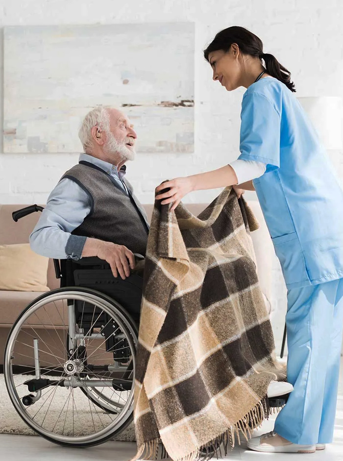 Nurse providing in-home support to an elderly man in a wheelchair by placing a blanket over him, demonstrating compassionate assistance and personalised care through NDIS home nursing care services.