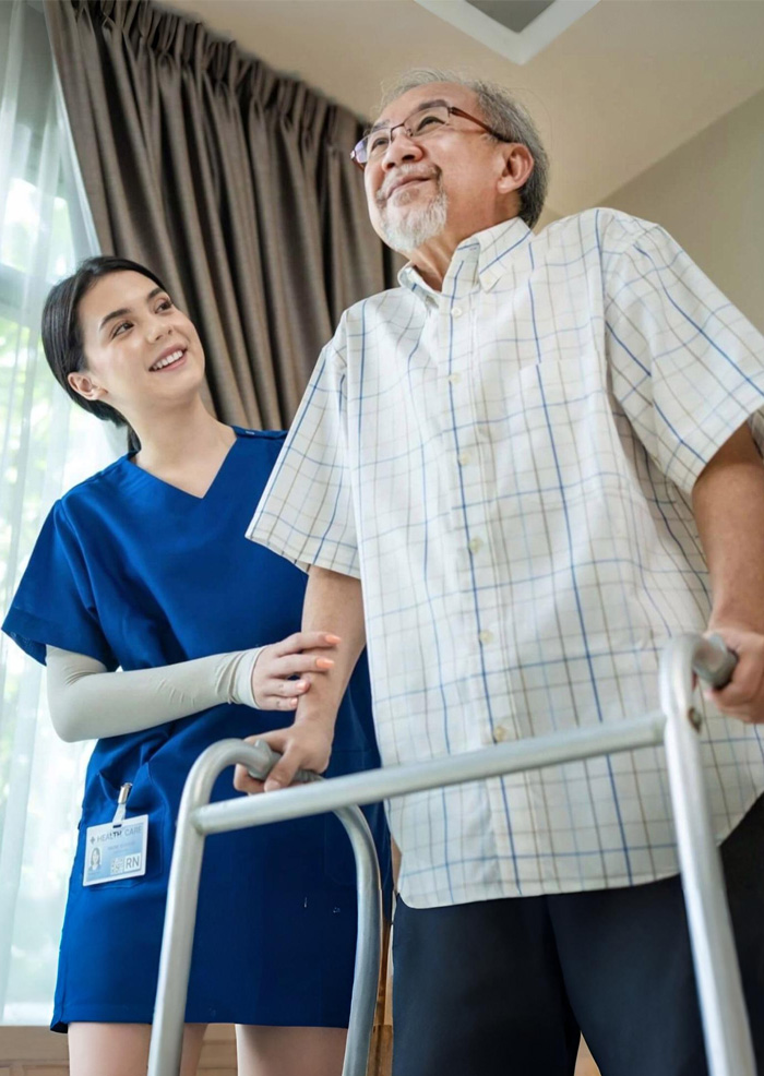 Nurse assisting an elderly man using a walking frame inside his home, demonstrating mobility support and personalised assistance provided through NDIS home nursing care services.
