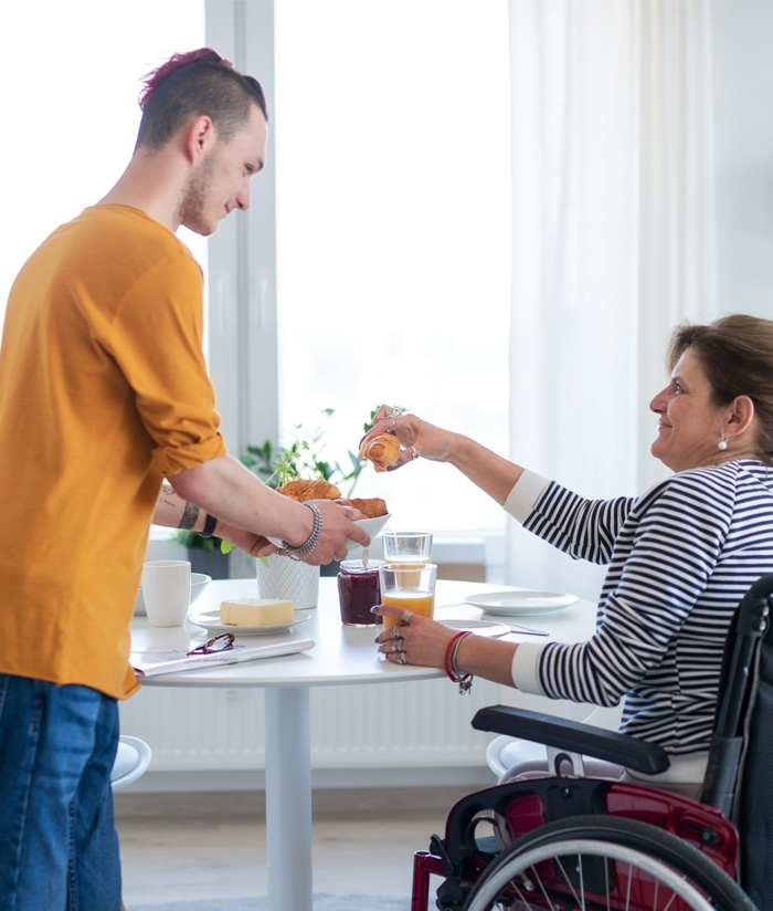 Support worker serving breakfast to a woman using a wheelchair at a dining table, demonstrating assistance with daily meals and independent living support through NDIS shared living services.