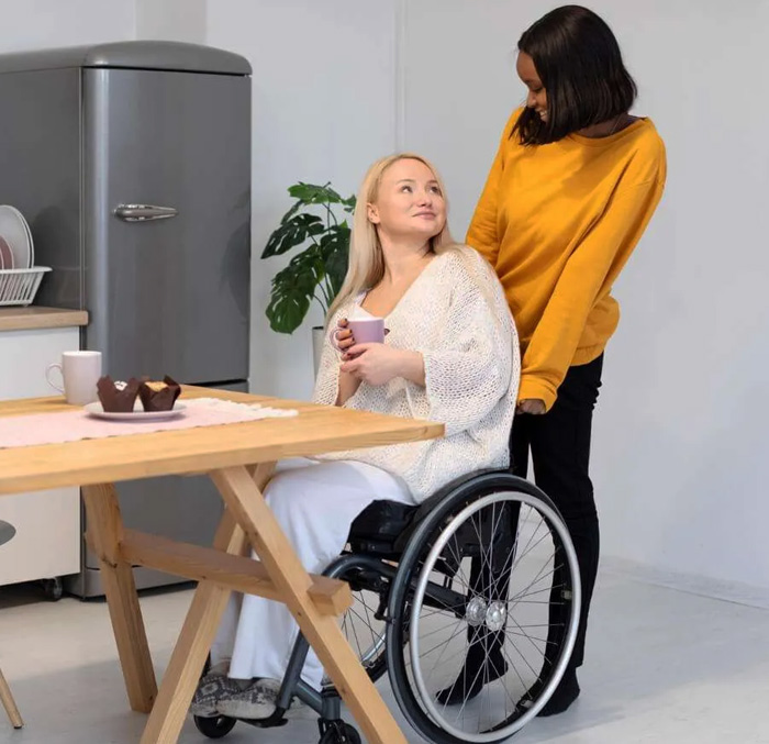 Support worker assisting a woman using a wheelchair in a kitchen while sharing a relaxed moment over coffee, representing personalised daily living assistance and supportive care in NDIS shared living services.