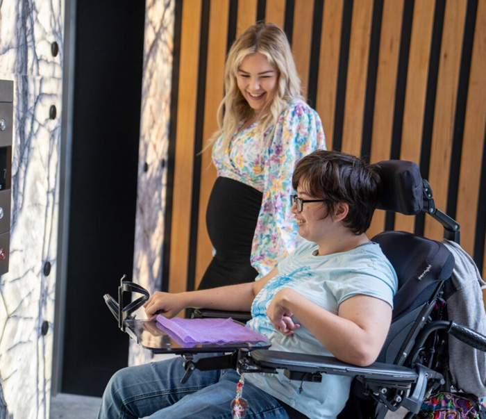 Support worker assisting a person using a powered wheelchair while operating an accessible lift control panel, illustrating independence, mobility support, and daily assistance provided through NDIS shared living services.
